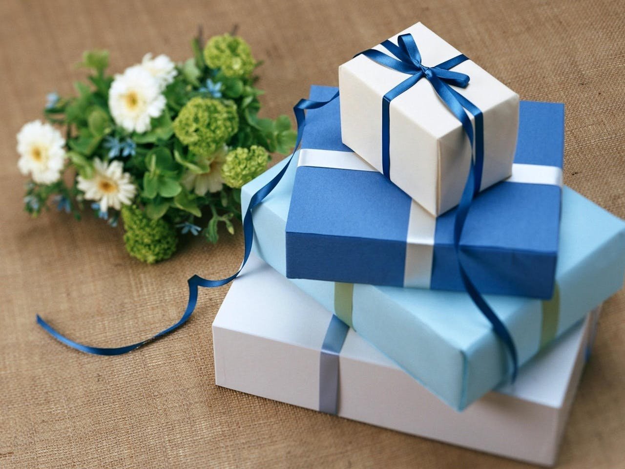 services-01 Three beautifully wrapped gift boxes with blue ribbons arranged beside a floral bouquet.