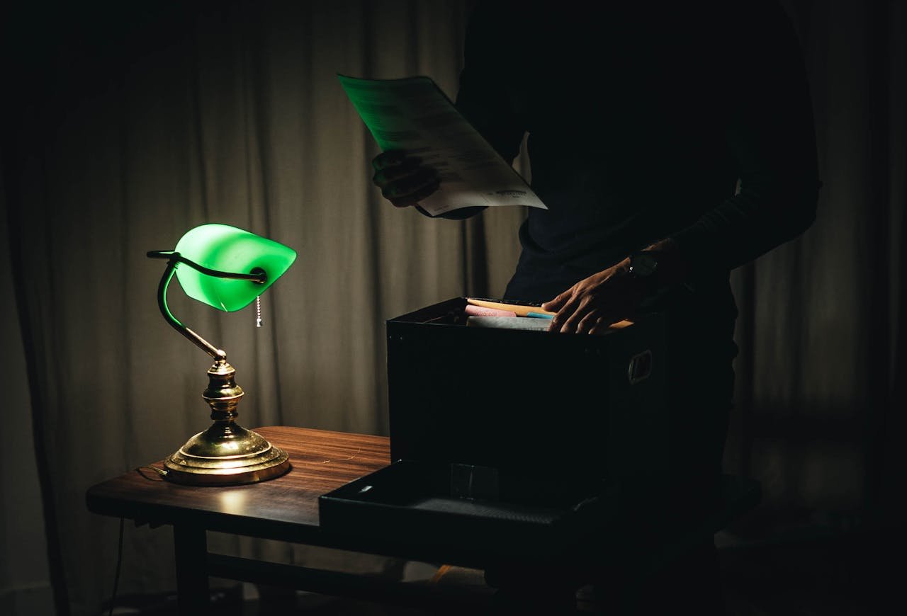 services-03 Unrecognizable male with sheets of paper standing near wooden table with glowing table lamp and box of documents in dark room while working late at home