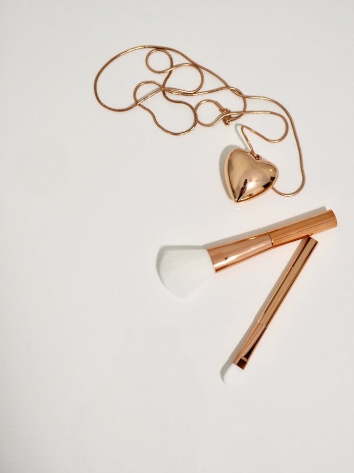 hero-img-01 A charming flat lay of a heart-shaped pendant and makeup brushes on a white background, creating a minimalist still life.